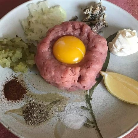Mound of hand-chopped beef tatar with a whole raw egg yolk nested in the centre, surrounded by separate piles of diced pickle, onion, paprika, pepper and salt with a lemon wedge