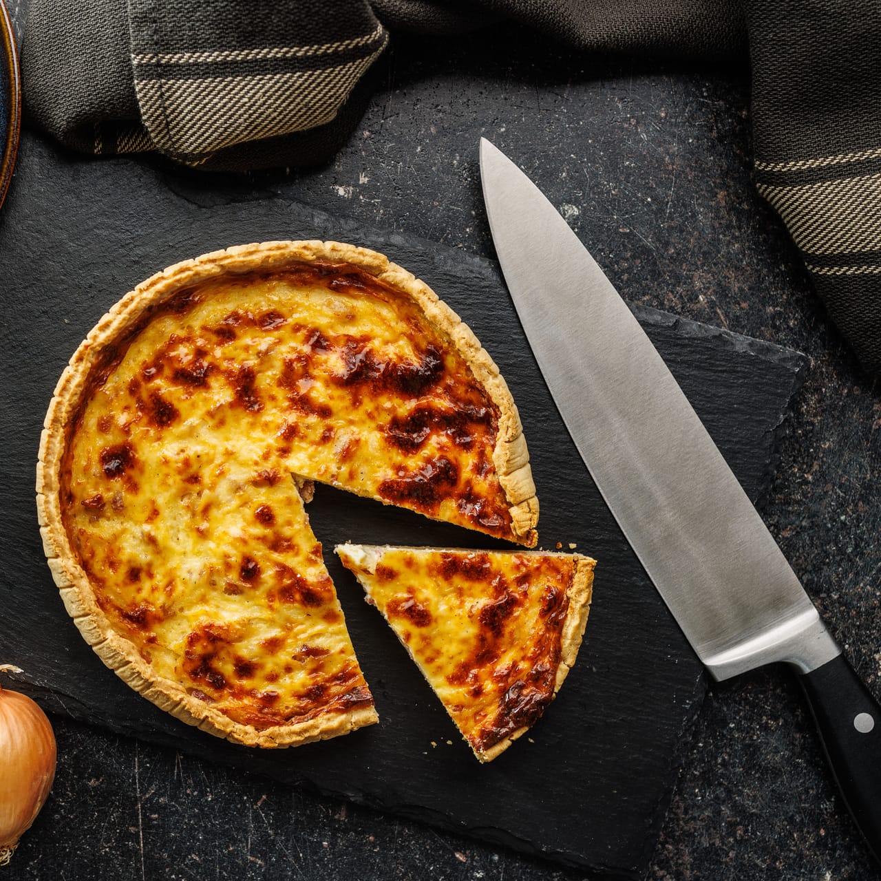 A sliced cheese quiche with a golden broiled top on a dark slate board, with a chef's knife and raw onions alongside