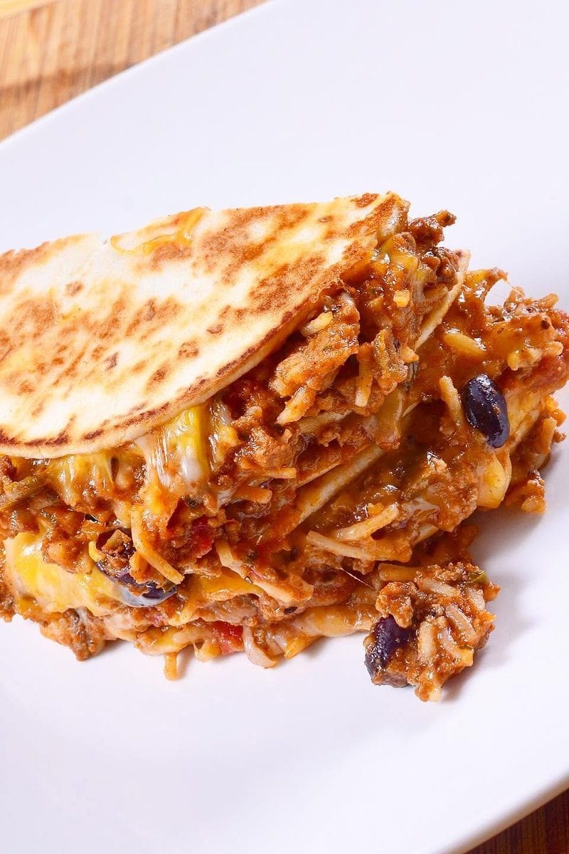 Cross-section of a stacked ground beef and Spanish rice quesadilla, its grilled tortilla layers oozing melted cheese and studded with black beans, on a white plate