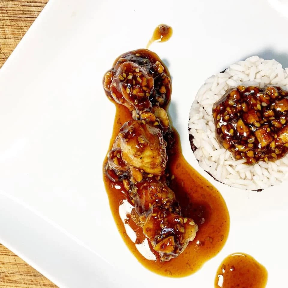 Glazed teriyaki pork bites glistening with sesame seeds beside a stacked mound of white and forbidden black rice, on a white plate