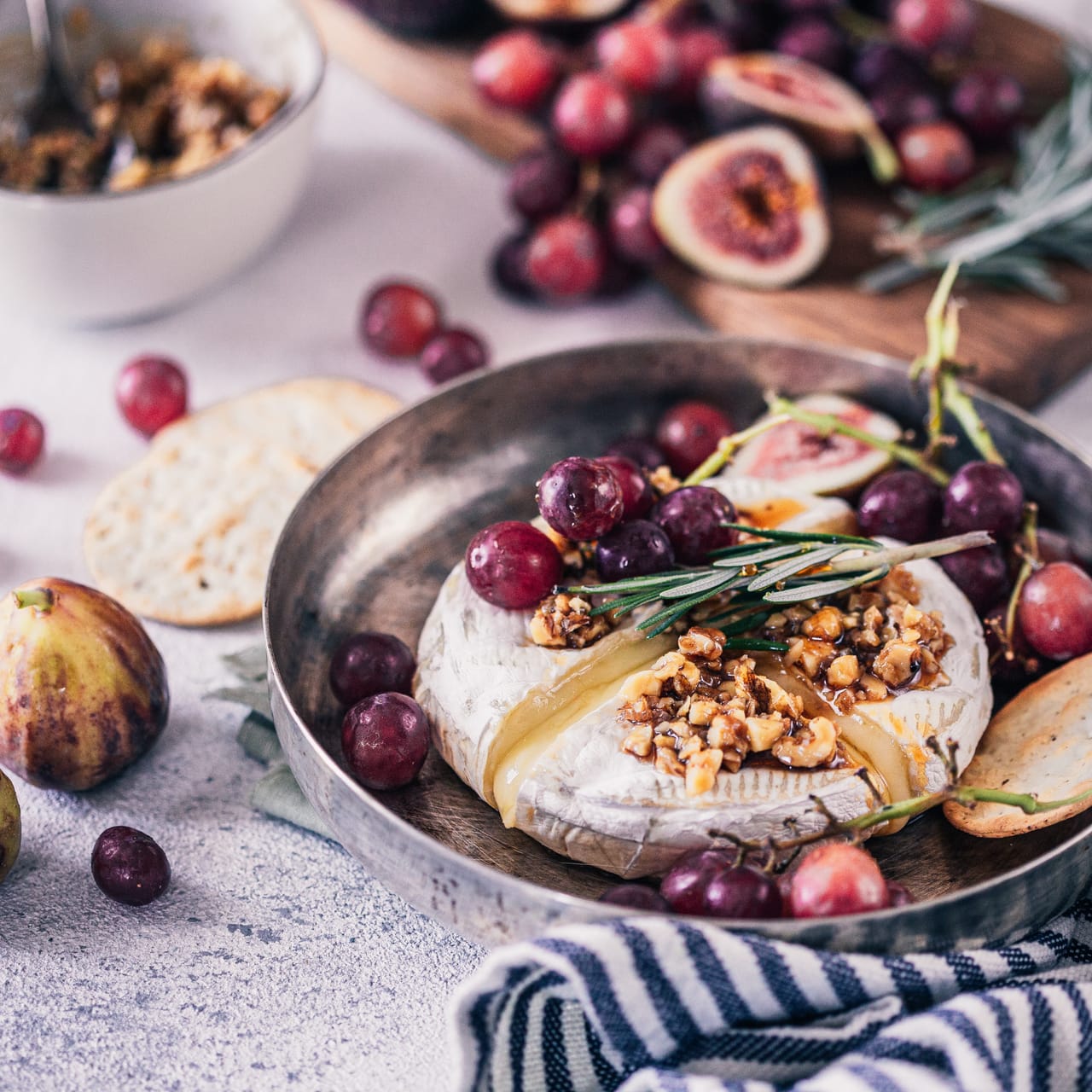 A wheel of baked brie topped with honey and chopped walnuts in a metal pan, surrounded by crackers, grapes, and figs