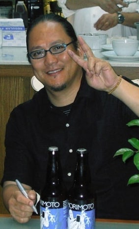 Masaharu Morimoto smiling and giving a peace sign, sake bottles on the counter in front of him