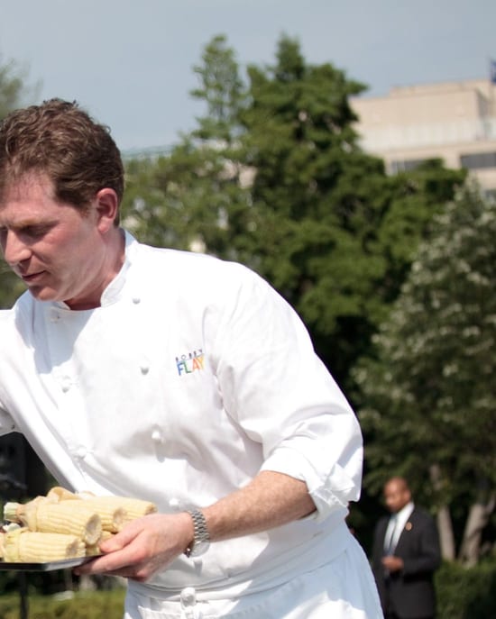 Bobby Flay at a grill in a white T-shirt and black cap, corn and steak on the grate