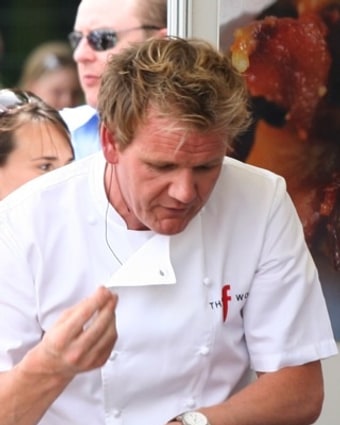 Gordon Ramsay in chef's whites, head down over a pan at an outdoor food festival, tasting from a spoon