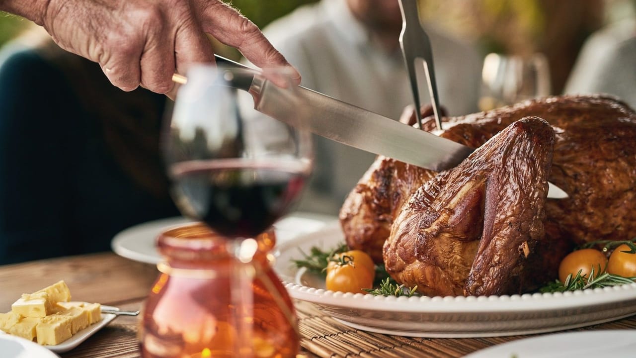 A hand slicing a roasted turkey leg with a carving knife and fork on a platter garnished with rosemary and cherry tomatoes