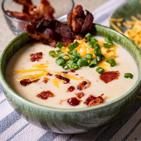 Green ceramic bowl of creamy potato soup topped with shredded cheddar, sliced green onions and crisp bacon chips